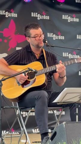 A man with glasses and dark hair plays an acoustic guitar while singing into a microphone. He is wearing a black shirt with a pattern and dark pants. The guitar is natural wood colored with a red pickguard. He is seated on a folding chair on a stage in front of a black backdrop with pink butterfly designs and the word "BAKGARDEN" repeated. There are stage lights visible above the backdrop. A music stand is positioned to his right.