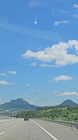A car drives down a highway under a bright blue sky dotted with fluffy white clouds. The sun is high, indicating it is daytime, likely midday or early afternoon. The landscape is mountainous, with rolling green hills covered in trees on either side of the road. In the distance, a town is visible at the base of a larger mountain with a distinct, rocky peak. The road appears to be a multi-lane highway with a metal guardrail separating the lanes. The overall scene suggests a scenic drive through a rural or semi-rural area. There are no people visible outside of the car. The mood is peaceful and pleasant, indicative of good weather for travel. No text is visible in the media.