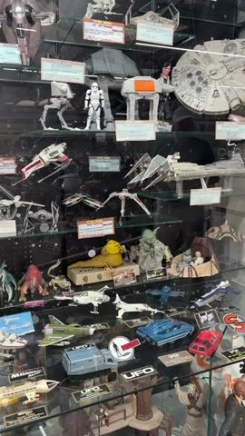 The video shows a display case filled with various models and action figures, primarily from the Star Wars franchise. The items are arranged on multiple shelves.

On the top shelf, there are several Star Wars vehicles, including what appears to be an AT-AT walker, a stormtrooper figure, and the Millennium Falcon.

The second shelf from the top features more Star Wars spacecraft, such as X-wings and other fighter-type vehicles.

The third shelf displays a collection of figures and smaller ships. Prominent characters include Jabba the Hutt and Yoda. There are also several alien-like figures and some smaller spacecraft.

The fourth shelf showcases a mix of models, including what looks like a space shuttle, a green fighter jet, and a red car-like vehicle. Some items have labels indicating "UFO" and "ALPHA MOONBASE."

The shelves below continue to display more models and figures, with a focus on science fiction themes. There are various spacecraft, including rockets and shuttle-like designs, as well as some alien creatures and characters. The display appears to be in a store or museum setting, with reflective glass in front of the items and a black background with a starfield pattern. The lighting is bright, illuminating the collection. No people are clearly visible in the immediate vicinity of the display.