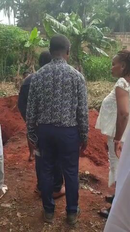 The video shows a group of people gathered around what appears to be a freshly dug hole in the ground. The ground is red clay soil, and the surrounding environment is lush with green vegetation, including trees and banana plants. The people are wearing a variety of clothing, ranging from casual to more formal attire. One person is seen holding a shovel near the hole. It is raining during the video, and it is possible that this gathering is related to a burial or a memorial service.