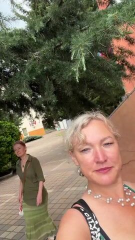 The video shows two women outdoors. The woman in the foreground, with blonde hair and wearing a pearl necklace, is looking towards the camera with a slight smile. She has her right arm raised behind her head. A woman with dark hair, wearing a green dress, is visible in the background, walking away from the camera. They are in an outdoor setting with trees and a paved walkway. Buildings are visible in the distance.