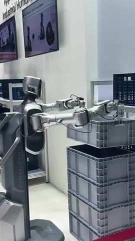 The video shows a humanoid robot in what appears to be an exhibition or demonstration setting. The robot has a metallic grey finish and is equipped with two articulated arms. One arm is extended and holding a grey plastic crate, which is being placed on top of a stack of similar crates. The robot is positioned on a mobile base, indicated by the wheels visible at its bottom. In the background, there are shelves with more crates, digital displays showing information about robots, and other exhibition elements like stanchions with red velvet ropes. The environment is well-lit and appears to be indoors.