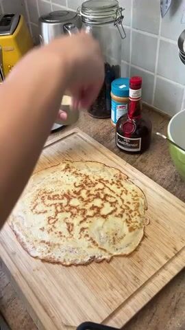 A person's hands are visible in the upper left corner of the frame. The person is holding a small, light-colored cup and appears to be sprinkling something onto a large, golden-brown crepe that lies flat on a wooden cutting board. The crepe has a slightly uneven, lacy edge and a patterned surface from cooking. In the background, on a kitchen counter, are several items: a jar filled with dark beans or coffee, a blue container, a jar of peanut butter, and a bottle of Grand Marnier liqueur. To the right of the crepe, a green bowl is partially visible. The scene is a close-up shot focused on the preparation of the crepe.