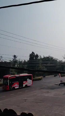 The video shows a view of a gas station and a street. On the street, a red and white bus is visible, and a motorcycle is riding away from the camera. The gas station has a large sign with the Pertamina logo and several fuel price indicators. There are several cars and motorcycles at the gas pumps. In the background, there are trees and a tall building. The sky is clear and bright. The camera appears to be stationary, looking down at the scene from a slight elevation.