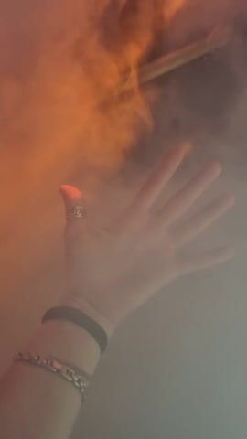 A hand with a ring on a finger is visible amidst low-lying fog or smoke. The fog obscures much of the background, but as the camera moves, people are seen walking through the smoky environment. The fog is illuminated by various colored lights, including red, orange, and teal. In the background, there appear to be lights strung up, adding to the festive atmosphere. The scene appears to be at a party or event with special effects.