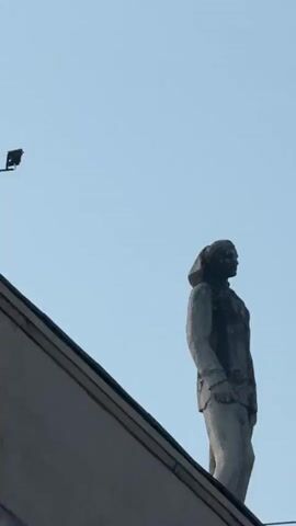 The video pans across a building's facade, focusing on statues adorning its roofline. The sky is a clear, pale blue. On the left, a surveillance camera is mounted on a thin pole extending from the building's edge. To the right, two stone statues stand atop the building. The one on the left appears to be a fragment of a larger structure, possibly a standing figure with an angular, broken top. The statue to its right is a more complete figure of a person in a long coat, facing to the right. Further to the right, another complete statue, also in a long coat, stands facing left. The building itself is made of concrete or stone, with ornate columns visible at the bottom of the frame. The overall impression is of an older architectural style, with elements of both classical design and possibly modernist additions like the surveillance camera.