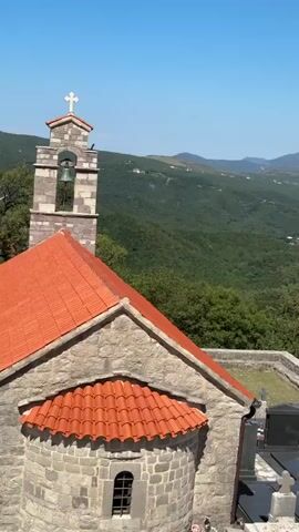 The video is a vertical panoramic shot of a hilly, green landscape under a clear blue sky. In the foreground, a stone building with a terracotta tiled roof is visible, possibly a church or a chapel, with a small section of a cemetery to its right. The cemetery has several tombstones, some with white crosses. The camera pans to the right, revealing a lush green valley dotted with buildings and trees, leading towards a body of water. In the distance, large mountains form a backdrop, their peaks partially obscured by haze. The overall impression is of a serene, natural landscape with a touch of human settlement.