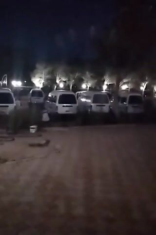 The video shows a nighttime scene with several white vans parked in an outdoor area. The ground appears to be dirt or gravel, with some scattered debris. In the background, there are trees and several lights illuminating the area. The vans are parked in a row, and some have their headlights on. The overall atmosphere is dark and somewhat deserted. There are no people visible in the video.
