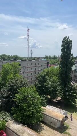 A high-angle drone shot pans right across a residential area on a sunny day. In the foreground, a red car is parked next to a series of corrugated metal sheds. Behind the sheds is a multi-story apartment building with rows of windows. Several tall, leafy trees surround the building. In the background, a tall telecommunications tower rises above the other structures against a partly cloudy blue sky.