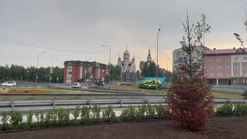 This media file captures an exterior view of a city street with a prominent Orthodox church in the background. The scene is a wide shot, showcasing a circular intersection with traffic flowing. Several buildings are visible, including a modern, multi-story building with a red facade on the left and a taller, pale building with multiple floors on the right. The church, with its golden domes, stands as a central architectural feature.

In the foreground, a small, reddish-brown coniferous tree is planted in a bed of dark soil, bordered by a line of green bushes. The road is divided by metal barriers and lined with what appear to be flowerbeds or landscaped areas. Cars are present on the road, indicating typical urban traffic.

The time of day appears to be overcast, possibly late afternoon or early morning, with a muted sky. The weather seems to be cloudy. The overall emotion conveyed is calm and ordinary, depicting a typical day in an urban environment.

There is some visible text in the media file. On the tall, pale building on the right, there is lettering that appears to be in Cyrillic, part of a name or sign. On one of the buildings on the left, a circular sign with red and white elements is visible, possibly indicating a traffic direction or information. There is also a blue and green triangular monument or sculpture in the middle ground, possibly a local emblem or landmark. The presence of the Orthodox church and Cyrillic text suggests the location is within a Russian-speaking region, consistent with Khanty-Mansiysk, Russia.