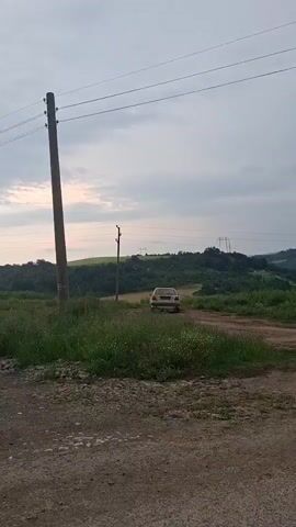 The video opens with a light-colored car driving away on a dirt road, receding into the distance amidst green fields and grassy areas. Two wooden utility poles with power lines are visible, with one in the foreground on the left. The background consists of rolling, tree-covered hills under an overcast sky.

The camera then pans to the right, revealing a panoramic view of a rural landscape near Balkantsi, Bulgaria. The dirt road transitions into an asphalt road in the foreground. A white, triangular road sign and a metal guardrail are visible beside the road. The view encompasses expansive green fields and hills stretching into the distance under the same cloudy sky.