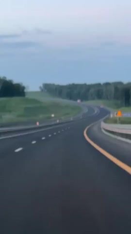 This is a first-person perspective video of a car driving on a highway. The highway is paved and has white dashed lines marking the lanes, with a yellow line separating the lanes on the right side of the screen. There are guardrails and orange traffic cones visible along the side of the road. The road curves to the left and then to the right. On either side of the highway are green grassy hills and a line of trees. The sky is a pale blue with some clouds, suggesting it is either early morning or late evening. There are no other vehicles or people clearly visible in the video. The overall impression is of an empty highway in a rural setting during twilight.