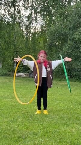 A young girl with pink-streaked hair is standing in a grassy yard in front of some trees. She is wearing a brown varsity jacket, a white top, black leggings, and yellow shoes. She has both arms outstretched, and she is twirling a yellow hula hoop with her right arm and a green hula hoop with her left arm. She is smiling at the camera.