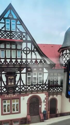 The video shows a close-up, slightly angled exterior view of a historic building with a red tiled roof and white walls. The building features distinctive dark brown half-timbering with intricate geometric patterns, including circles and diagonal lines. Several windows with white frames are visible across the facade. A sign above the main entrance reads "POSTAMT" in dark letters. The main entrance has a dark arched doorway and steps leading up to it. To the right of the entrance, a blue mailbox stands on the sidewalk. The sky is cloudy. The image is captured from a slightly elevated perspective, looking up at the building. The lighting suggests it is daytime.