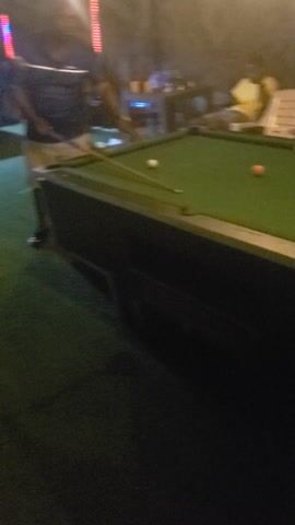 The video captures a lively nighttime scene, likely within a bar or recreation center, where people are playing pool and socializing. The lighting is generally dim, with bright spotlights illuminating the green-felt pool tables, creating a high-contrast atmosphere. The video is shot vertically and appears handheld, with frequent panning and slight movements.

At the 0:00 mark, a man wearing a dark shirt with white stripes is seen leaning over a green pool table, holding a cue stick, and preparing to hit a white cue ball. Other blurred figures are visible in the background. Throughout the video, upbeat music with a distinct beat and vocals plays in the background.

From 0:01 to 0:02, the camera pans to the right, revealing more of the establishment. Another green pool table comes into view, where a person in a light-colored (possibly yellow) shirt is taking a shot. The background is dark, but flashes of red and blue lighting suggest a bar or club setting.

Between 0:02 and 0:03, the camera continues to pan, showing multiple groups of people gathered around at least two pool tables. Individuals are standing, sitting, and observing the games. A green object ball is visible on one of the tables.

The scene remains similar from 0:03 to 0:04, emphasizing the social aspect of the gathering. An orange object ball is now visible on one of the tables.

By 0:04 to 0:05, the camera refocuses on a man (possibly the same one from the beginning or the one in the yellow shirt) leaning over a pool table, cueing up a shot with a white cue ball aimed at an orange object ball. A green floor covering, resembling turf or carpet, is visible in the lower part of the frame. The overall ambiance is casual and energetic, indicative of friends enjoying a night out playing pool.