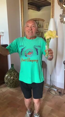 An elderly bald white man wearing a bright green t-shirt and black shorts stands in what appears to be a room with terracotta tile flooring. He is holding a clear glass vase with yellow flowers in his right hand, and his left hand is raised with fingers spread. The t-shirt has a graphic on it that includes the words "BIG GAME" and "Best Company" above and below an image of a fishing lure. In the background, to his left, is a large green glass bottle filled with what appears to be dried herbs or flowers. To his right is a tall, white floor lamp and a large ornate mirror. The man is smiling and appears to be speaking to the camera.
