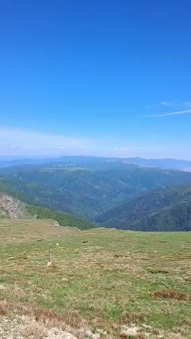 The video presents a panoramic view of a mountainous landscape under a clear, blue sky. In the foreground, there is a grassy field with patches of reddish-brown vegetation. Beyond the field, a range of green, tree-covered hills and mountains stretches into the distance. Further back, a row of windmills is faintly visible on a distant ridge. As the camera pans from left to right, the view reveals rocky terrain and a higher mountain with snow-covered patches and craggy peaks. The overall scene evokes a sense of altitude and expansive natural beauty.