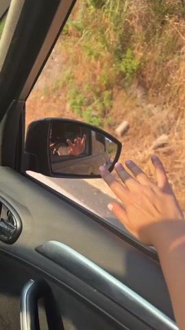 A woman's hand, with manicured nails, rests on the exterior of a car door, touching the side mirror. In the reflection of the side mirror, a woman is visible inside the car, holding a phone and making a peace sign with her left hand, while her right hand is raised and blurred as she waves or gestures. The background outside the car is a dry, hilly landscape with sparse vegetation, illuminated by sunlight. A sign is visible in the background of the mirror reflection with text that appears to read "DOBRO DOŠLI WELCOME" and "OPŠTINA BUDVA MUNICIPALITY OF BUDVA". The car interior, visible around the door and mirror, consists of dark gray and silver trim.