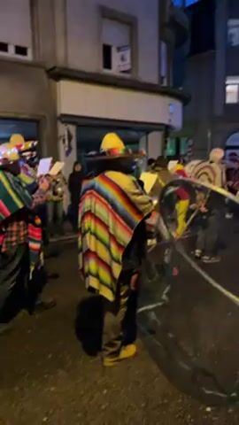 The video depicts a nighttime street scene with a group of people dressed in colorful clothing, ponchos, and sombreros. They appear to be part of a parade or celebration, moving down the street.  A small cart with drums and other percussion instruments is being pushed by one of the individuals. Some spectators watch from the sidewalks. The street is illuminated by streetlights and building lights, giving the scene a vibrant and festive atmosphere.