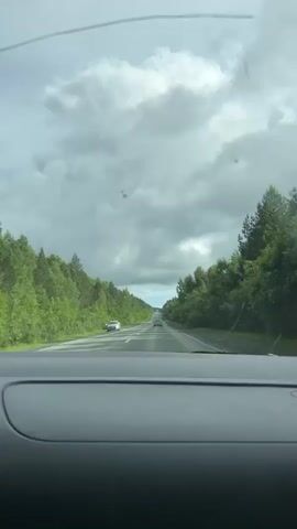 The video is a first-person perspective from inside a car looking out the windshield. The car is driving on a two-lane highway with a grassy median. Dense green trees line both sides of the road, and the sky is overcast with gray clouds. A white car is parked on the shoulder to the left, and several other cars are visible in the distance ahead. The road appears wet, suggesting recent rain.