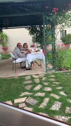 A man and a woman are seated at a table under a green gazebo. The table is covered with a white tablecloth and set with plates, glasses, and bottles. The man is wearing a light-colored checkered shirt and white pants. The woman is wearing a patterned dress. They appear to be eating and interacting. The gazebo is surrounded by grass and paved areas. Red roses are blooming on the side of the gazebo. In the foreground, there are large, flat stones placed on the grass. The background shows a building and some potted plants. The video is slightly blurry and appears to be filmed from a distance.