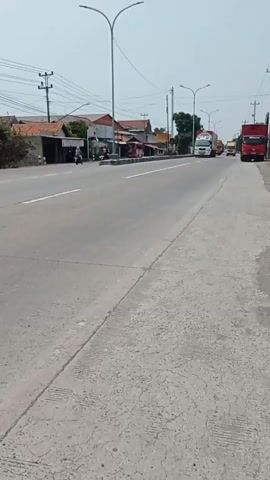 The video shows a street scene with several trucks. In the foreground, a concrete sidewalk is visible with some cracks and tire marks. In the background, a paved road stretches into the distance. Several trucks are driving on the road. On the left side of the road, there are buildings and houses with red tile roofs. There are also utility poles with power lines. The sky is clear and bright. A motorcycle is seen passing by on the left side of the road.