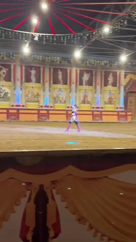 The media file shows a person dressed in a costume, possibly a gladiator or a warrior, performing in an arena. The performer is wearing a metallic gold and black outfit with pink boots and leg coverings, and a crown-like headpiece. They are holding a blue prop, which appears to be a sword or a staff, and are in various dynamic poses, including standing and kneeling. The arena has a sandy floor and is decorated with backdrops that resemble ancient Roman architecture, with columns and murals depicting historical scenes and statues. Blue lights are strategically placed, illuminating parts of the arena. The overall atmosphere suggests a live performance or show, possibly a circus or a themed event. There is no visible text in the media file. The location context provided, Kamchatka, Russia, does not appear to be reflected in the visual elements of the media file, which strongly suggests a Roman-themed setting.