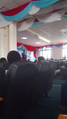 The video shows a lecture hall filled with rows of black, cushioned chairs occupied by people. A man stands in front of the room, likely giving a presentation. The hall is decorated with red, white, and light blue draping fabric across the ceiling. There are windows on the right side of the room. A few people are standing by the entrance. The floor is covered with a blue carpet. The lighting appears to be standard fluorescent.
