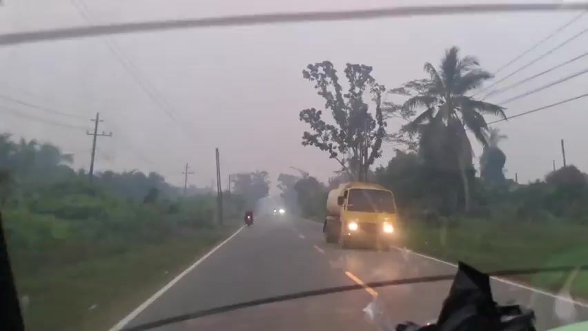The video shows a view from inside a vehicle, looking out through the windshield. It appears to be a cloudy or hazy day, possibly early morning or late afternoon, as the light is dim. The vehicle is on a paved road, with green vegetation and trees lining both sides. There are utility poles with wires visible along the roadside. Several motorcycles are on the road, some moving in the same direction as the camera vehicle and others in the opposite direction. One motorcycle in the foreground appears to have a rider and is moving away from the camera. Further down the road, two headlights from an approaching vehicle are visible. On the right side of the road, a yellow tank truck is partially visible, also moving in the same direction. The overall impression is of a rural or semi-rural road with ongoing traffic.