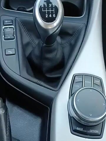 The video shows the interior of a car, focusing on the dashboard and center console. The car has a manual transmission with a six-speed gear shift. There are buttons for sport and eco pro modes, as well as parking assist. The infotainment system displays music information and navigation. The climate control settings are visible, with the temperature set to 19.5 degrees Celsius. The video then shifts to show the view from the driver's seat, looking out onto a street with houses and a white van parked in the distance. The speedometer and other gauges are visible.