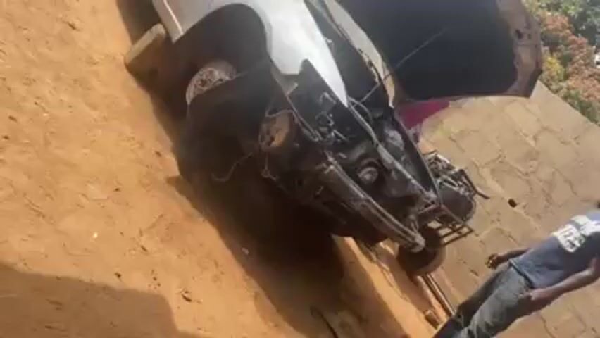 The video shows a partially disassembled white car on a dirt lot. The car's hood is open, revealing the engine, and the front tires are missing. The car is propped up on blocks.  Several people are visible around the vehicle, moving and appearing to be working on or inspecting it. The background shows a wall and some trees.  The scene appears to be a repair or salvage area.