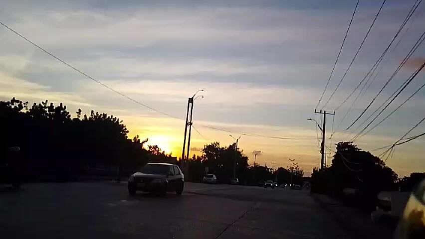 The video shows a road at sunset. There are several cars driving on the road. The sky is a mix of orange, yellow, and blue colors. There are trees and power lines on either side of the road.