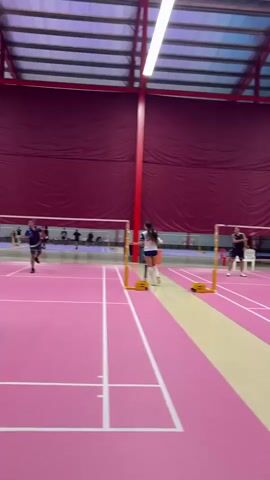 A badminton match is underway indoors. A female player in a white and blue uniform with white socks and sneakers stands on the right side of the court. She holds a badminton racket in her right hand and is looking to her left. Another player, a male in a blue and black shirt and dark shorts, holding a racket, stands on the left side of the court. He is looking towards the female player. In the background, several other people are present, some of whom appear to be playing or observing. The court has a pink surface with white lines. The background features a deep red curtain-like material hanging from the ceiling. The net of the badminton court is visible between the two players.