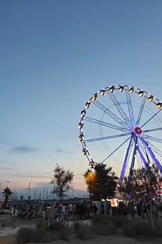 The video shows a Ferris wheel at dusk. The sky is clear with a slight orange glow near the horizon, indicating sunset. The Ferris wheel is illuminated with blue and white lights, and many of its gondolas are visible. Below the Ferris wheel, there is a crowd of people, some standing and some walking. There are trees and some structures, possibly stalls or ticket booths, surrounding the base of the Ferris wheel. The ground appears to be a mix of sand and paved areas. In the background, the water can be seen, with a few boats docked. The overall atmosphere is that of an amusement park or a fairground at the end of the day.