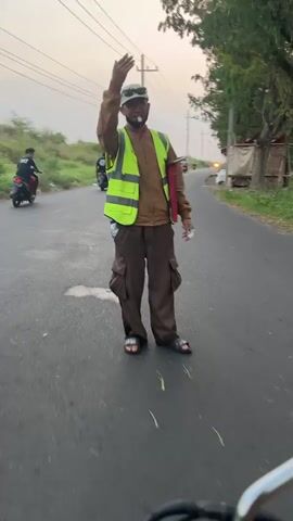 The video shows a man standing in the middle of a paved road, wearing a light brown long-sleeved shirt, dark brown cargo pants, a high-visibility yellow vest, a baseball cap, and sandals. He is waving his right hand. He is holding a red folder or binder in his left hand. Behind him, a motorcycle with two people on it is moving away. To the left of the road, there is vegetation and a dirt path. To the right, there are trees and a structure that appears to be a small building or hut. The sky is overcast, suggesting either dawn or dusk. The road has some puddles, indicating recent rain. The overall environment suggests a rural or semi-rural setting.