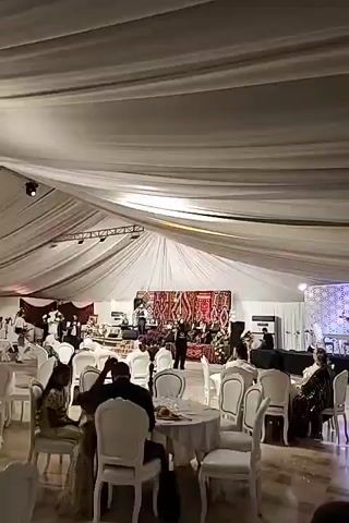 A video displays a wedding reception inside a large tent with white draping. In the center, a stage is set up with a backdrop of colorful woven rugs. A band is performing, with a male singer in the foreground. Several musicians are playing instruments, including drums and a keyboard. Guests are seated at round tables with white tablecloths and chairs, some of whom are watching the performance. The lighting appears to be dim, creating a festive atmosphere.
