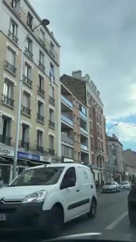 The video shows a street in a European town.  Buildings line both sides of the street, varying in height and architectural style. A white van is parked on the left side of the street. Further down, a yellow van is also parked. Several other cars are visible. A pedestrian is seen walking across the street. The sky is partly cloudy. The camera appears to be positioned from inside a vehicle. There are various store signs visible on the buildings.