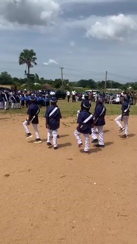 A group of children in matching navy blue uniforms with white trim and white pants march in formation on a dirt field. They are all wearing matching hats. In the background, there are more children and adults watching. There are also some cars and trees visible. The sky is partly cloudy.