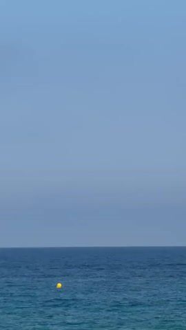 The video displays a tranquil ocean scene with a clear, pale blue sky dominating the upper portion of the frame. A distinct horizontal line separates the sky from the deep blue ocean, which occupies the lower half. A single, bright yellow buoy is visible in the water, positioned slightly to the left of the center and appearing to bob gently on the surface. The water itself is a mix of deep blue and lighter turquoise hues, with subtle ripples indicating calm conditions. No people or other objects are present in the frame. The overall impression is one of vastness and serenity, with the buoy serving as the sole focal point on the water.