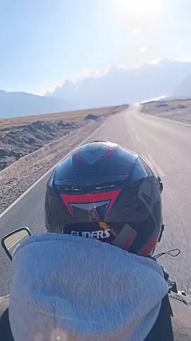 A first-person perspective video shows someone riding a motorcycle on a paved road through a mountainous landscape. The rider is wearing a black and red helmet with "GLIDERS" written on it, and a gray hoodie. The sun is shining brightly in a clear blue sky. The road stretches ahead, curving slightly to the right. On either side of the road are barren, rocky hills. In the distance, snow-capped mountains form a majestic backdrop. The video is short, lasting only four seconds, and appears to be filmed during the daytime.