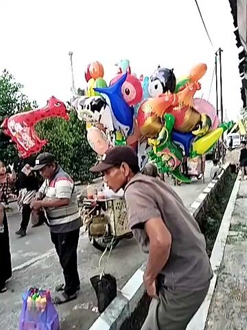 A person in a hijab and another person stand by a pink balloon shaped like a dinosaur and a collection of colorful balloons. A man in a striped shirt and a cap stands near them, holding a small object. Another man in a grey shirt and a cap is partially visible in the foreground. To the right, a red ice cream cart with an umbrella is visible, with various cartoon characters depicted on it. A green ice cream cart with text on it is to the left, next to a motorcycle. In the background, there are buildings and trees. The scene appears to be an outdoor market or street.