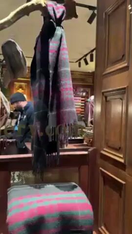 The camera peeks through a wooden door frame into what appears to be a shop interior.  A Scottish-style scarf hangs in the foreground obscuring some of the view.  A male employee, bald with a beard and glasses, is working behind a counter. He is wearing a brown vest over a white shirt and a bow tie.  Behind him is a shelf stocked with merchandise.  The shop is decorated with dark wood paneling and track lighting.