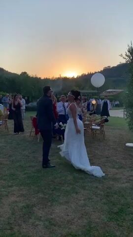 A wedding reception is underway outdoors as the sun sets. A man in a dark suit and a woman in a white wedding dress are dancing in the foreground. Several guests are visible in the background, some clapping and cheering. The reception appears to be set in a grassy area with chairs and tables set up. A large white balloon is also visible in the sky. The lighting suggests it is early evening.