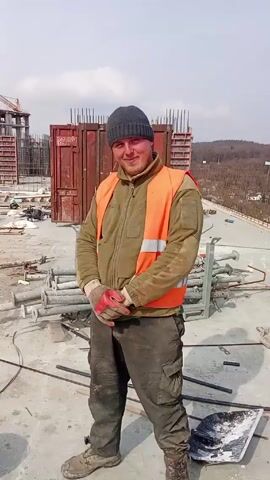 On a construction site on a bright day, a man wearing a grey beanie, an orange safety vest, a green fleece jacket, brown pants, work gloves, and dirty work shoes stands smiling at the camera. Behind him are construction materials, including metal forms, pipes, and rebar on a concrete surface. A building structure with a crane in the background is visible, along with some distant trees. There are some clouds in the sky.