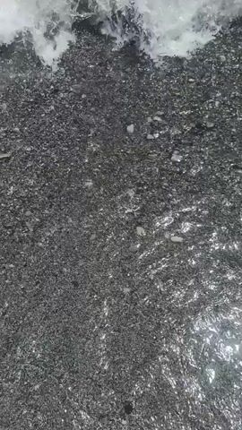 The video shows a first-person view of feet wearing flip-flops standing on a dark, wet surface. The surface appears to be composed of small pebbles or gravel. Waves are seen coming in and washing over the feet. The water is foamy and white as it hits the shore. The overall impression is of standing at the edge of the ocean or a lake.