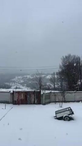 A snowy day. There is a patch of snow covered ground and an old rusty metal fence. There is also a small, white trailer sitting in the snow. Behind the fence is a slightly snow-covered hill and bare trees. Snowflakes fall from a gray sky.