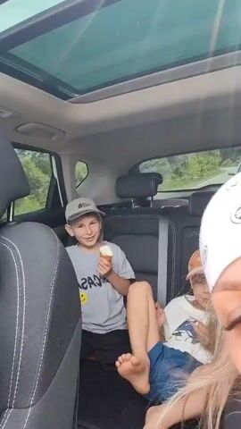 The video shows two young boys sitting in the back seat of a car. The boy on the left, wearing a grey t-shirt, blue shorts, and a grey baseball cap, is holding an ice cream cone and smiling at the camera. His legs are crossed, and his bare feet are visible. The boy on the right, wearing a white t-shirt, blue shorts, and an orange baseball cap, is also holding an ice cream cone and appears to be eating it. The interior of the car features black seats with white stitching and a panoramic sunroof. Outside the car, through the windows, a green, tree-covered landscape is visible, suggesting the car is in a rural or natural setting. The video appears to be shot from the front passenger seat, with the arm and shoulder of a person, possibly the driver, visible in the lower right corner. The lighting is bright, indicating it is daytime.