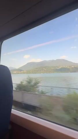The video shows a view from a train window looking out at a large body of water, possibly a lake, with mountains in the background. The water is rippled and reflects the blue sky. There are some white clouds in the sky. The mountains are covered in trees and some buildings are visible on the hillsides. The train appears to be moving, as evidenced by the slight blur of the trees and fencing visible outside the window. A dark blue seatback is visible on the left side of the frame, suggesting the perspective is from inside a train carriage.