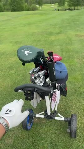 The video shows a golfer on a golf course. Initially, a hand wearing a white golf glove is on the handle of a white push cart holding a set of golf clubs. The camera then shifts to a selfie perspective, revealing the golfer smiling at the camera. Two other men are also visible on the green, walking towards the camera. The golfer appears to be walking alongside them, capturing the scene as they stroll across the lush green golf course under a cloudy sky.