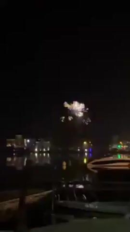 The video begins at night with fireworks exploding over a body of water. Buildings with lights are visible in the background, as well as boats. The camera then shifts to a man with long brown hair and glasses who is smiling. He is wearing a plaid shirt and a white turtleneck. The camera shifts again to a blurry shot of lights in the distance.